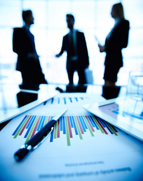 Technological devices and financial document with pen at workplace on background of three business partners communicating